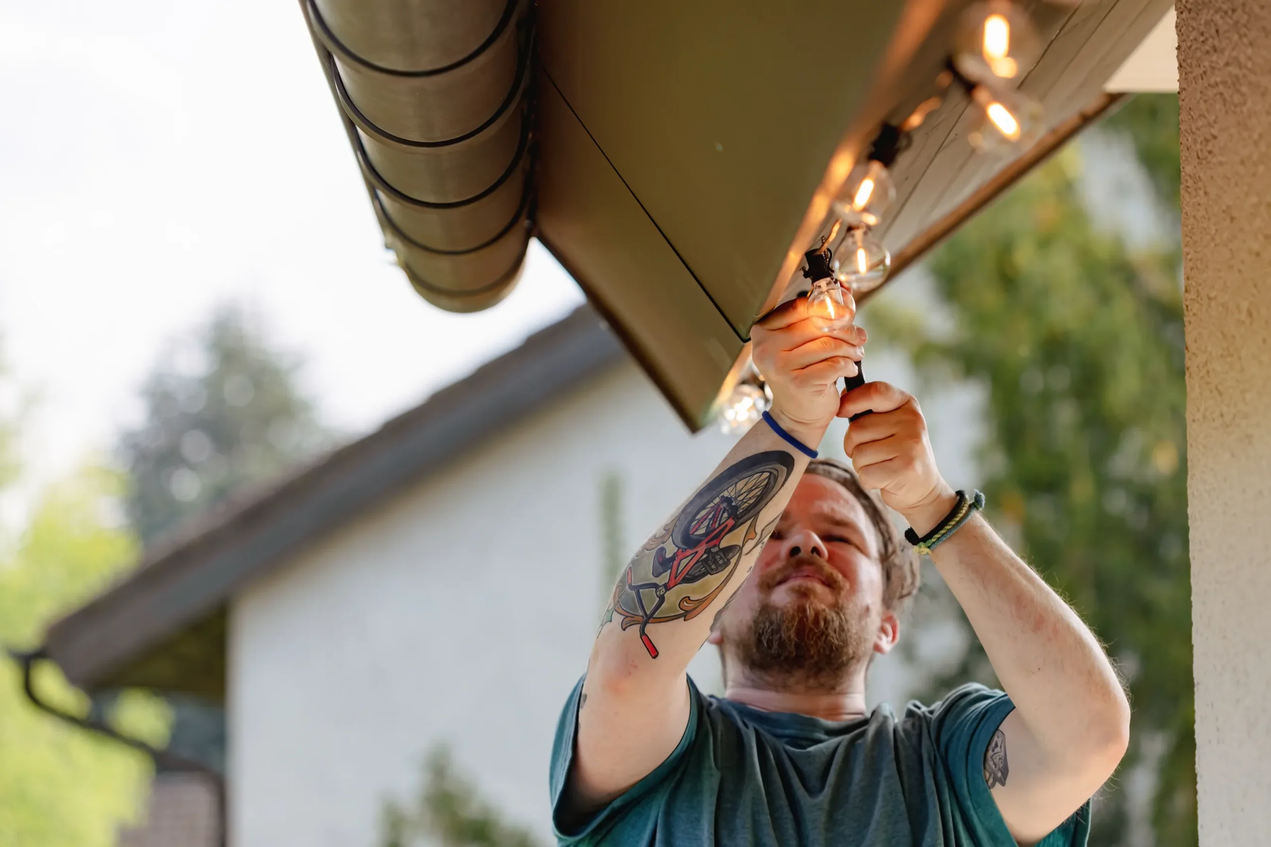A person with arm tattoos hangs string lights under a house eave, concentrating intently. The background shows a blurred green garden and adjacent houses.