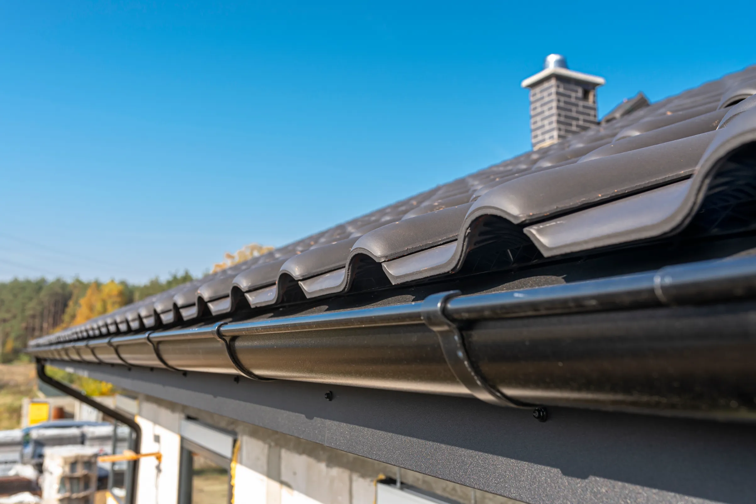 Close-up of a modern house's black tiled roof with a visible chimney and gutter system against a clear blue sky, conveying durability and elegance.