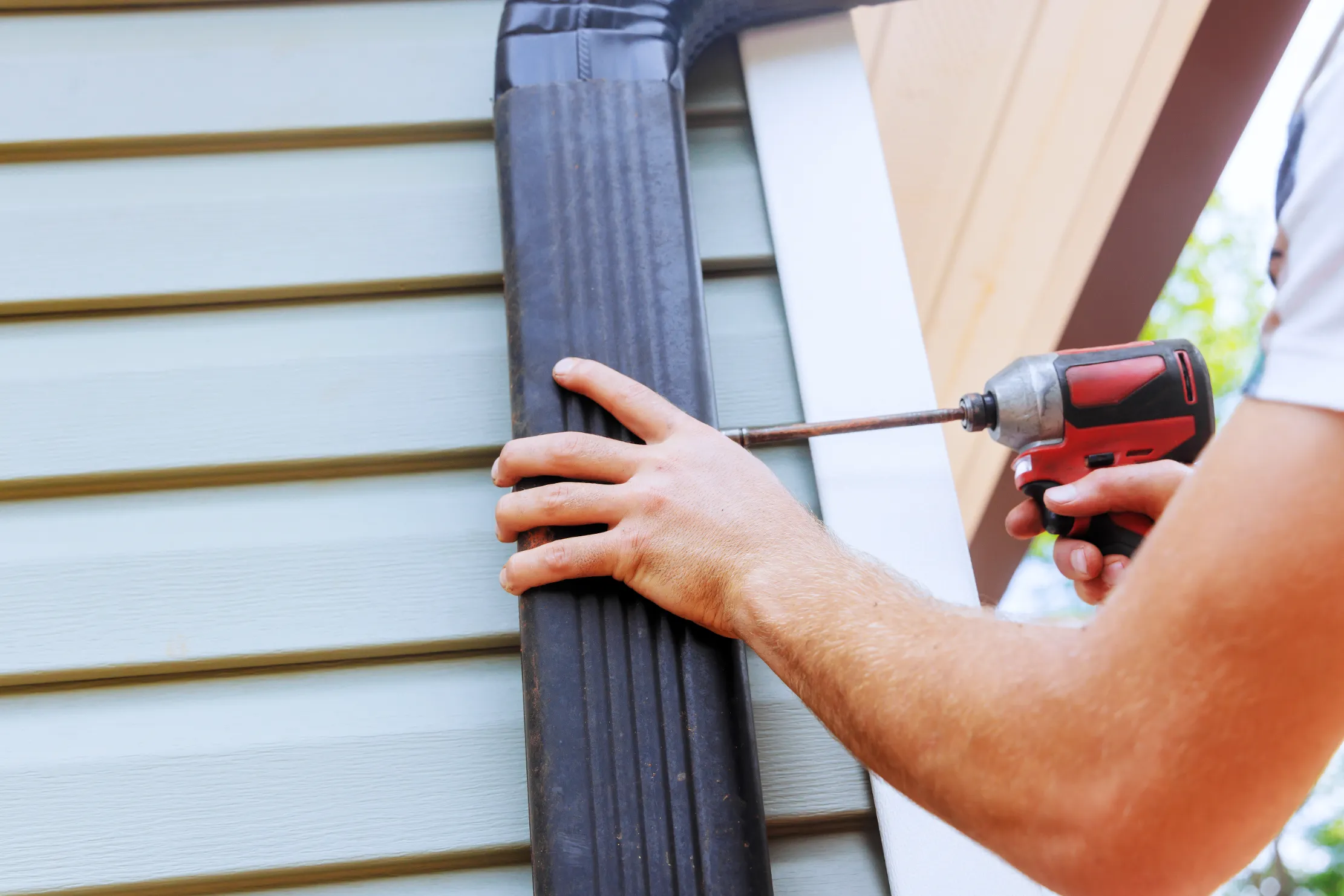 A person using a drill to attach a black downspout to the exterior wall of a house with light green siding, conveying focus and precision in home improvement.