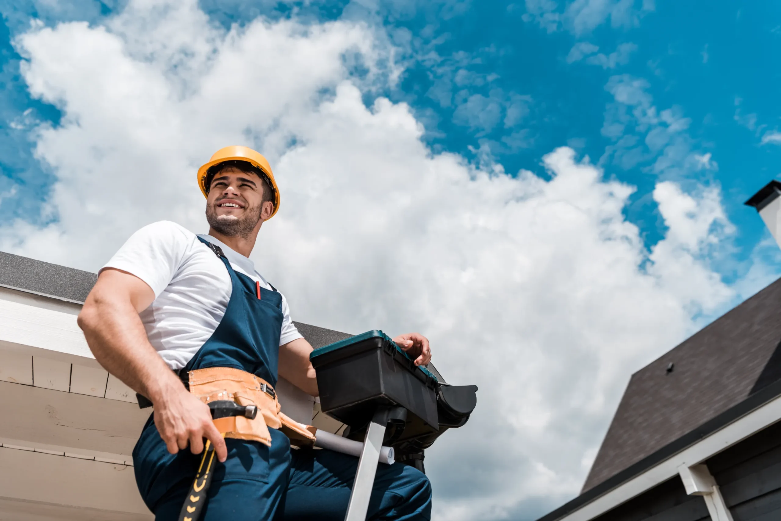 A smiling construction worker in a hard hat stands on a ladder under a cloudy blue sky, holding a toolbox and a tool belt, conveying confidence.