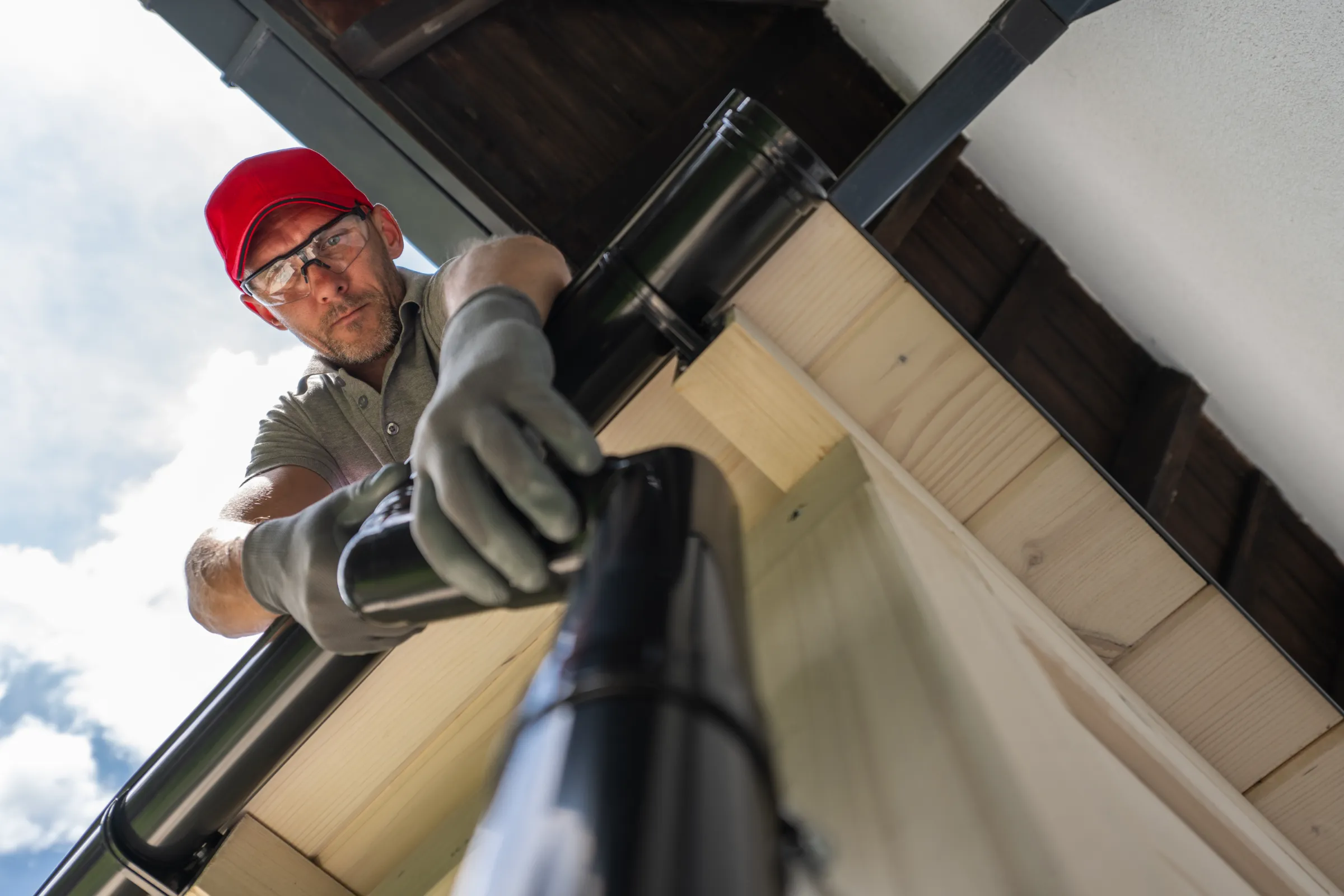 Man in a red cap and safety glasses, wearing gloves, installs a black gutter on a wooden roof. Sky with clouds visible, suggesting outdoor work.