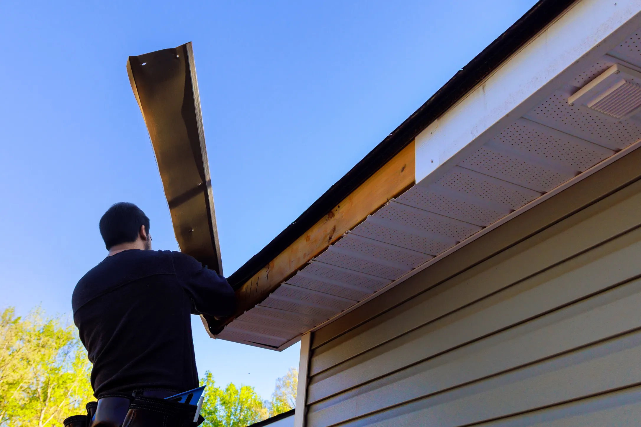 A person repairs the eaves of a house, lifting a metal piece under a clear blue sky. The scene conveys a sense of focus and outdoor work.